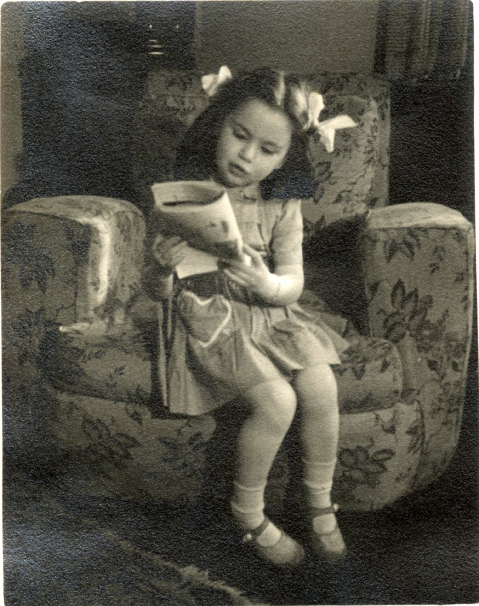 Me (with more bows in hair) sitting in a big armchair with a magazine in my hands,1945